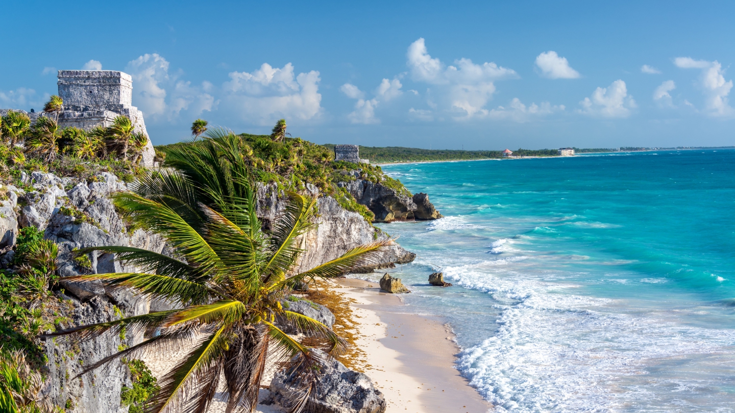 Scenic view of ancient Mayan ruins perched on a cliff overlooking the turquoise Caribbean Sea in Tulum, Mexico, with palm trees swaying along the shoreline.