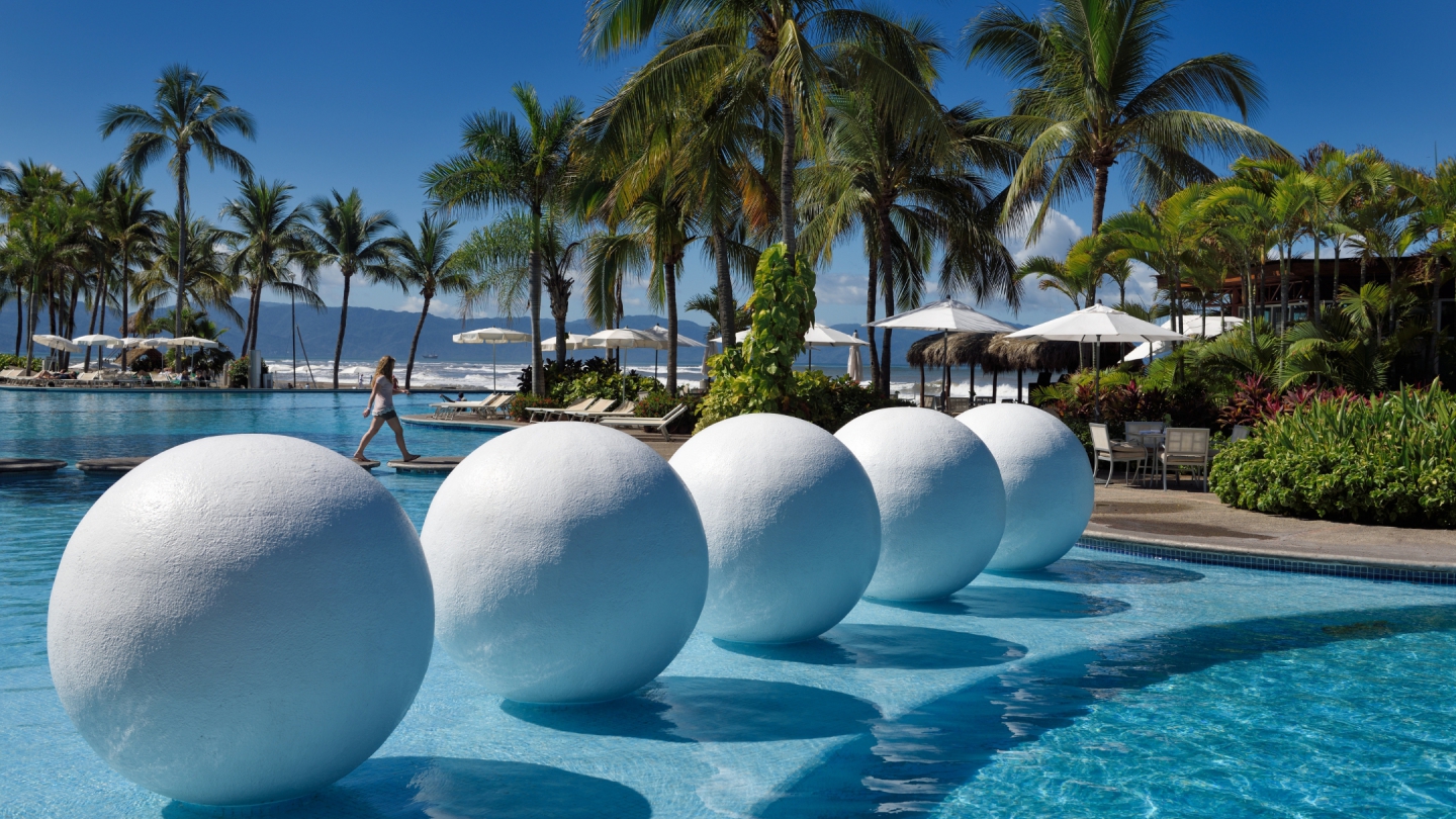 Modern beachfront resort pool in Mexico with palm trees, oversized white pool sculptures, and lounge chairs set against the Pacific Ocean horizon.