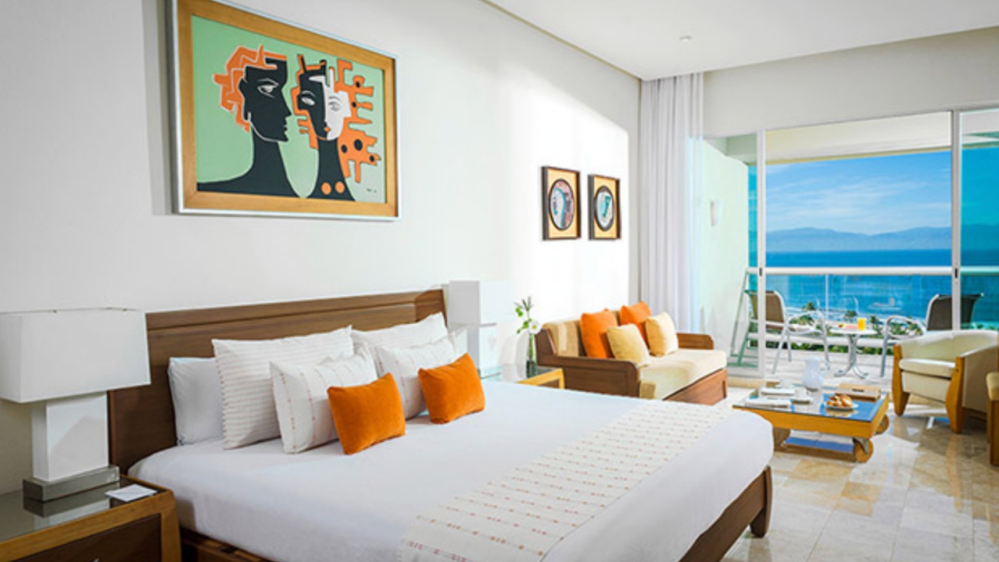 Ocean-view hotel suite in Mexico featuring modern decor, a private balcony, and a cozy bed with orange accent pillows overlooking Banderas Bay.