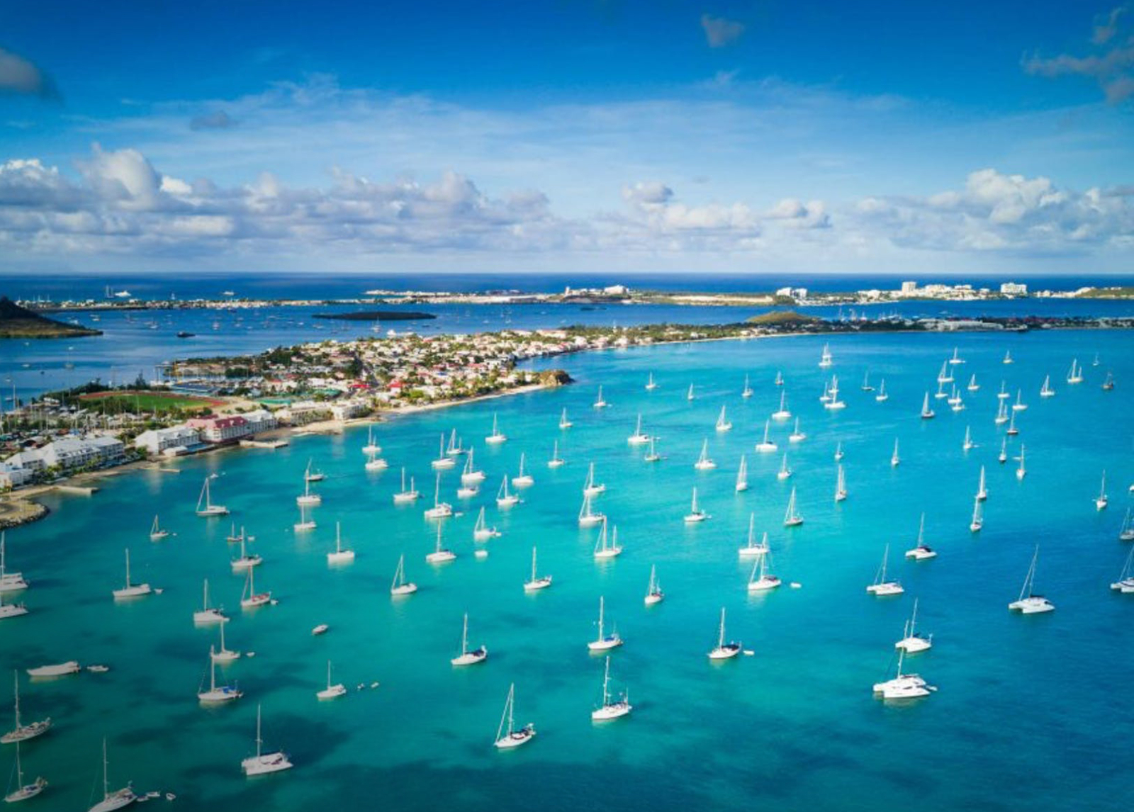 Aerial view of St. Martin’s turquoise harbor filled with dozens of anchored sailboats, with coastal homes and lush island terrain stretching into the distance.