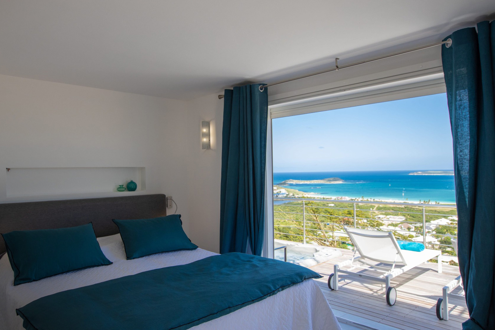 Bright villa bedroom with teal accents and floor-to-ceiling windows offering panoramic views of St. Martin’s coastline and ocean horizon from a private balcony.