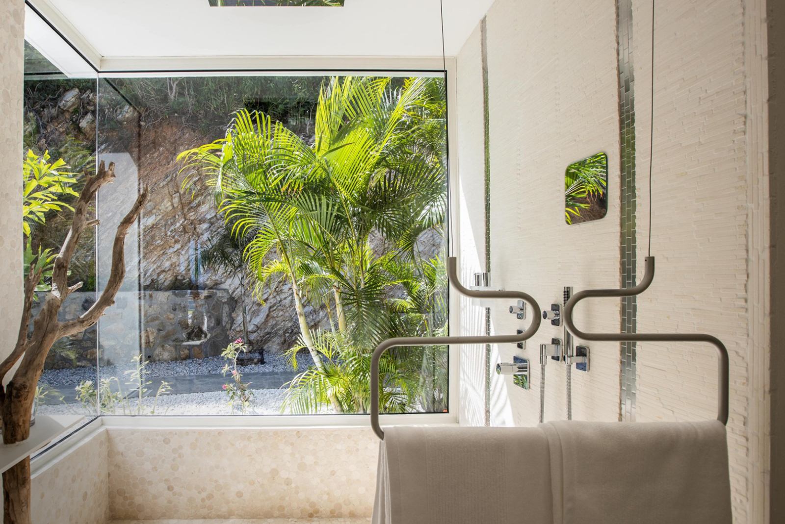 Modern indoor-outdoor bathroom in St. Martin with a glass wall showcasing tropical greenery, elegant stone tiling, and contemporary rainfall shower fixtures.