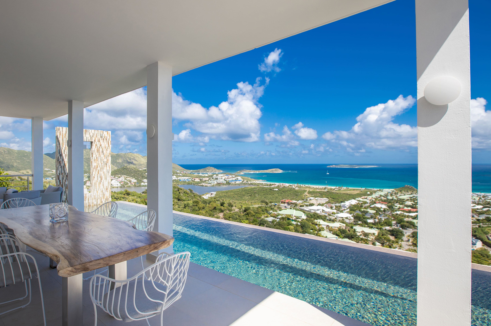 Covered outdoor dining area with a natural wood table, modern chairs, and a stunning infinity pool overlooking the turquoise Caribbean Sea and lush hills of St. Martin.
