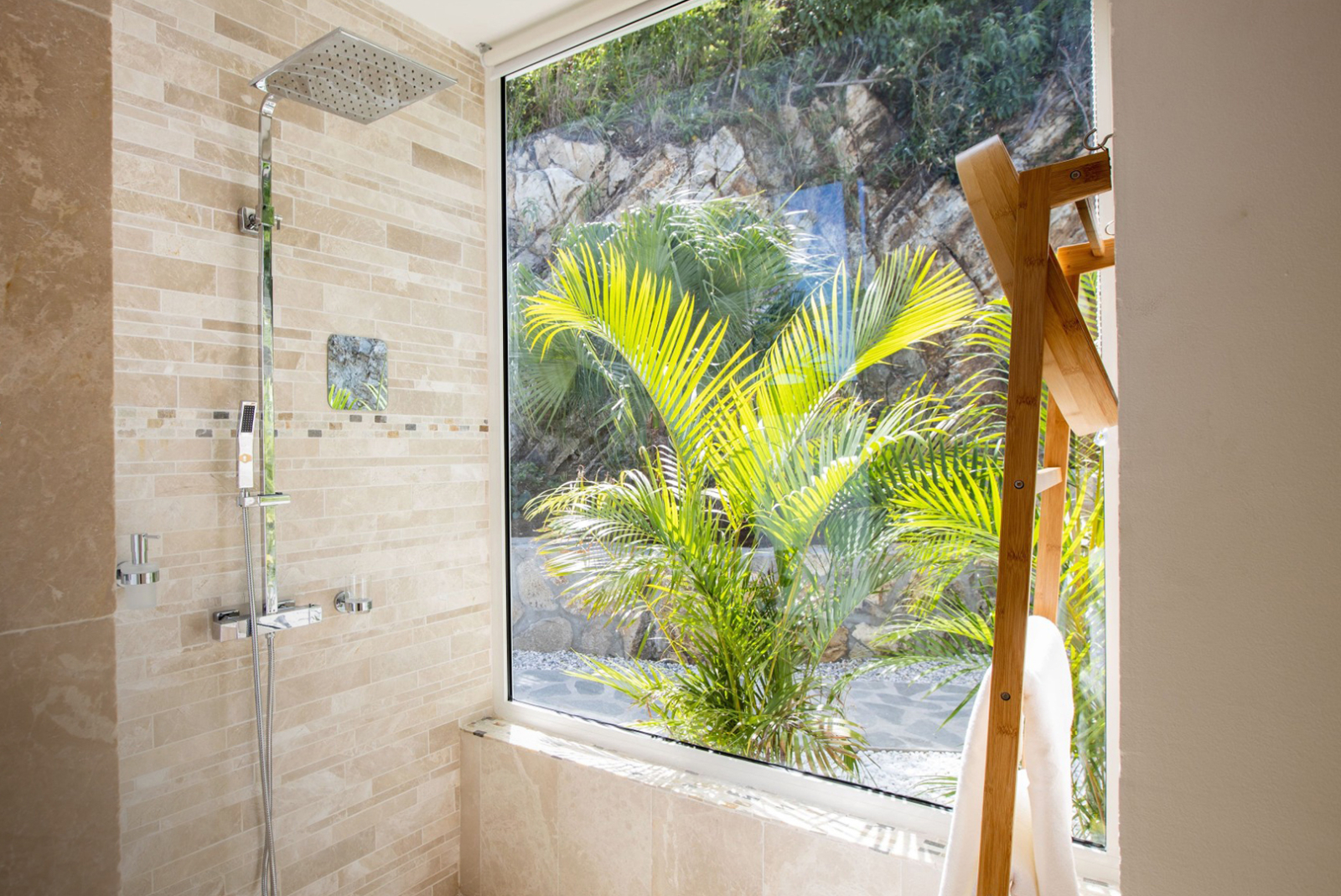 Luxurious tiled shower with a large window offering views of tropical plants and a natural stone wall, blending indoor comfort with outdoor serenity.