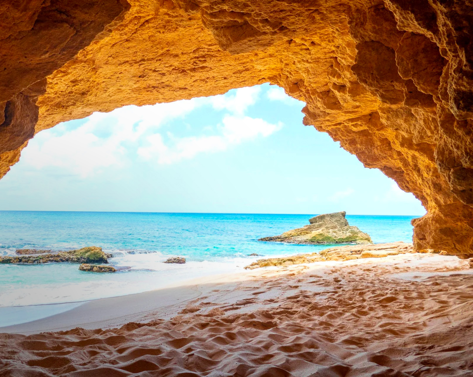 Golden sandstone cave framing a secluded beach and gentle waves, with a rocky islet in the distance under a soft, sunny sky.