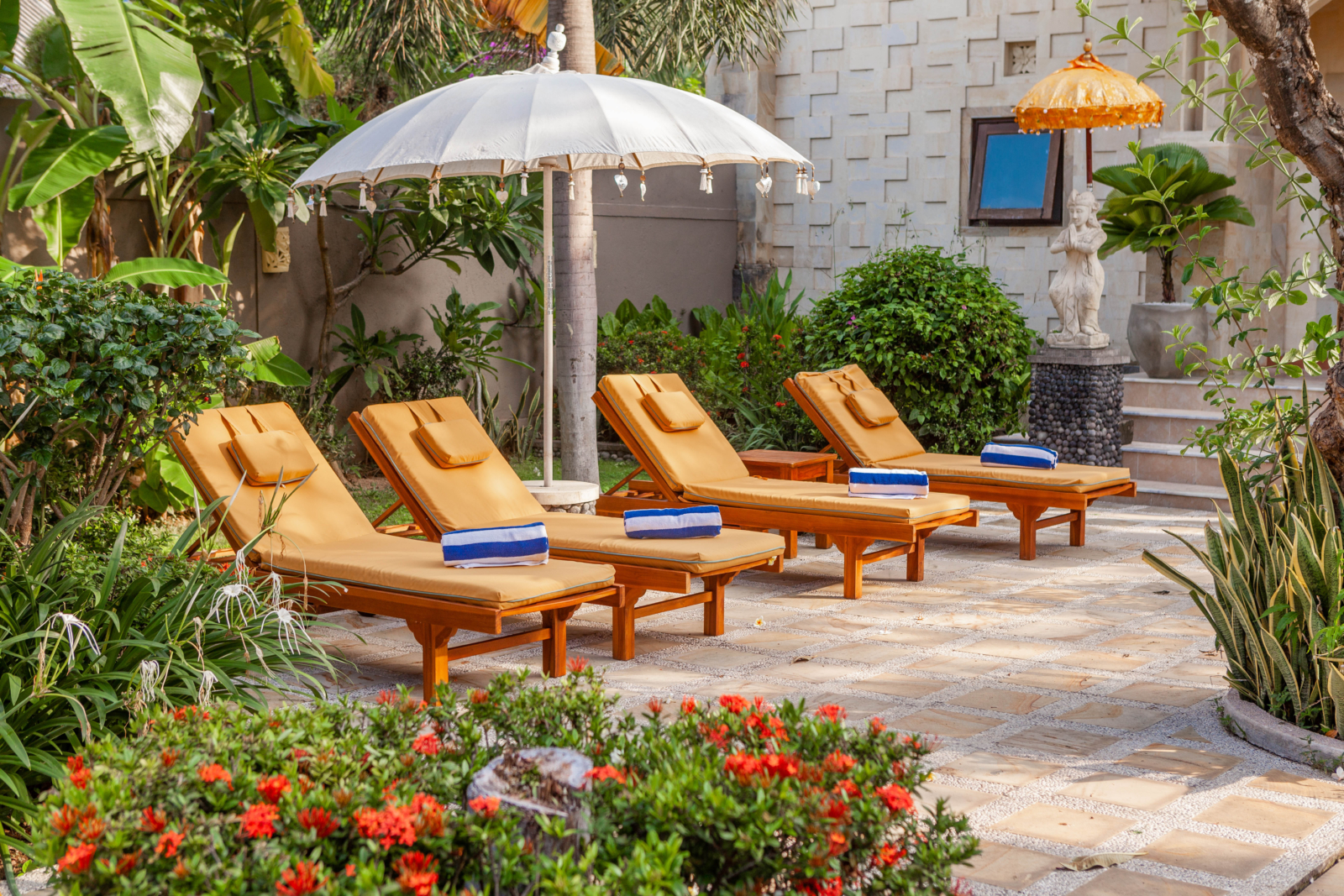 Relaxing outdoor lounge chairs by a sunny pool with vibrant plants and traditional Balinese decor.